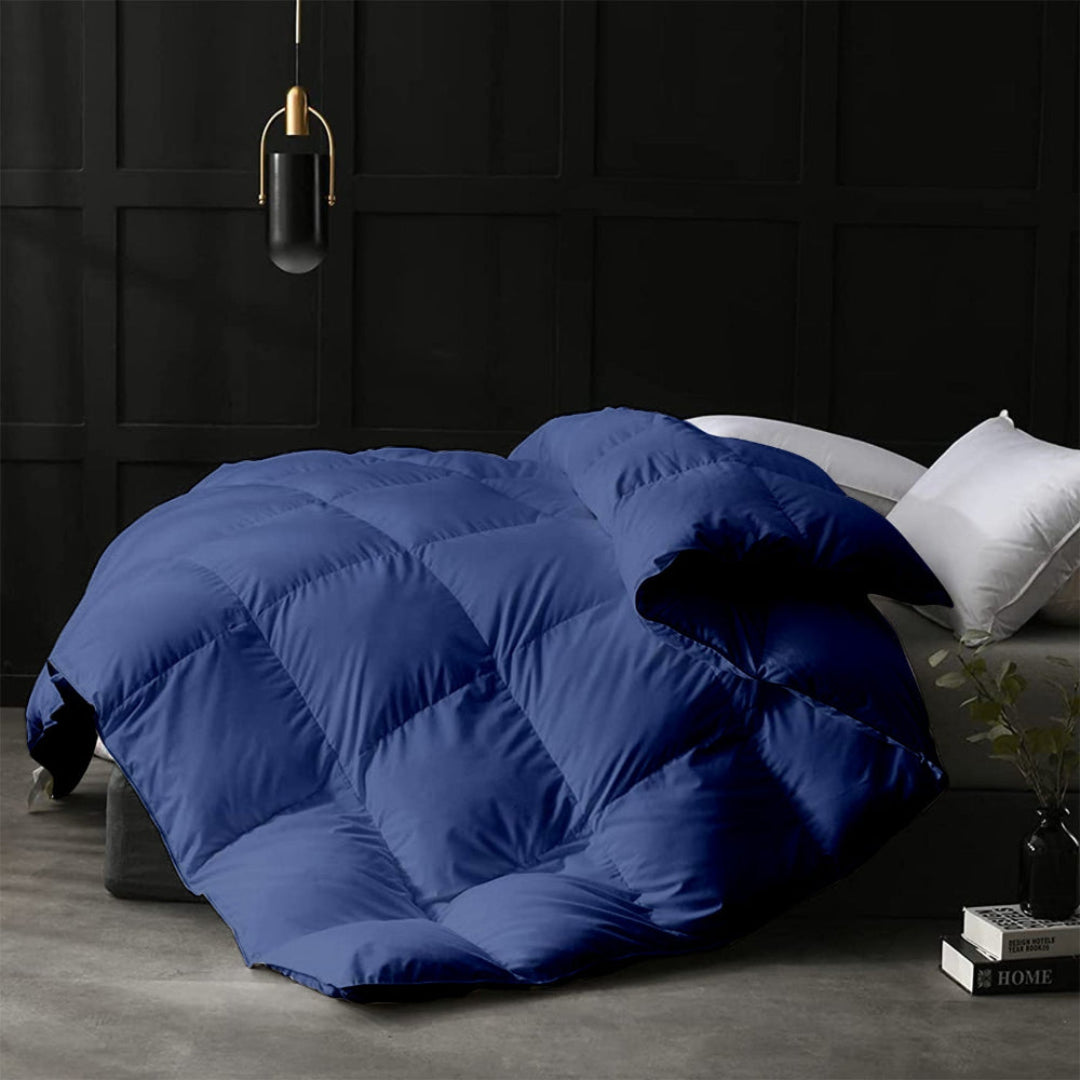 Blue quilt on a bed in a darkly decorated room with a lamp and books.