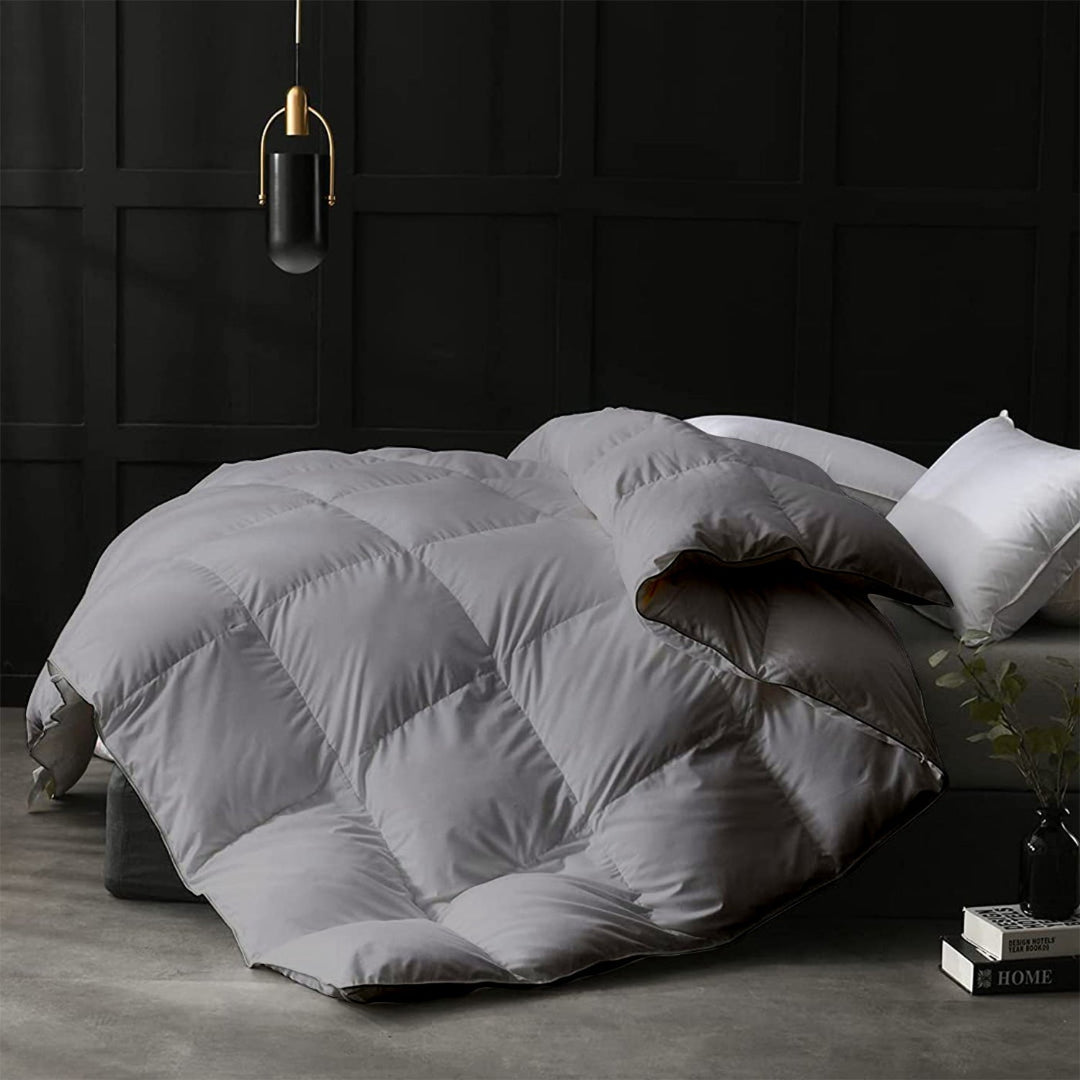 Gray quilt on a bed in a darkly decorated room with a lamp and books.